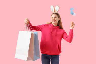 Alışveriş torbaları ve pembe arka planda kağıt tavşanlı tavşan kulaklı genç bir kadın. Paskalya kutlaması