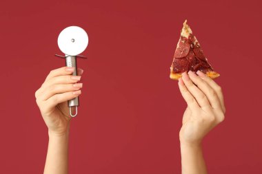 Kırmızı arka planda pizza dilimi ve pizza kesicisi tutan kadın eller.