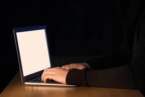 Hacker using laptop at table on dark background, closeup