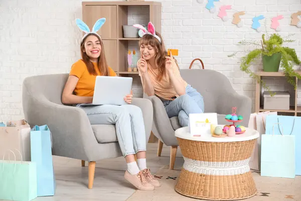 Happy young women in bunny ears headbands with laptop, credit card and paper bags for Easter at home. Online shopping