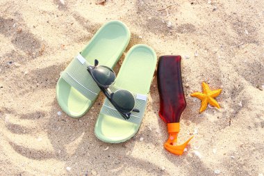 Şık güneş gözlükleri, güneş kremi ve kumlu arka planda parmak arası terlikler.