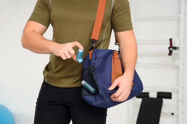 Sporty young man with bag and inhaler in gym, closeup