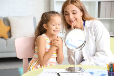 Mature speech therapist working with little girl near mirror at table in office