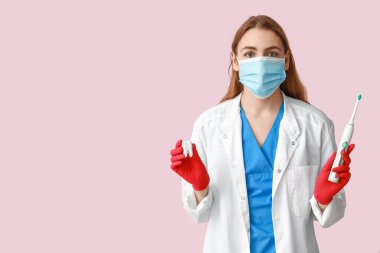 Maskeli, elektrikli diş fırçası ve pembe arka planda diş modeli olan kadın bir dişçi. Dünya Diş Hekimi Günü