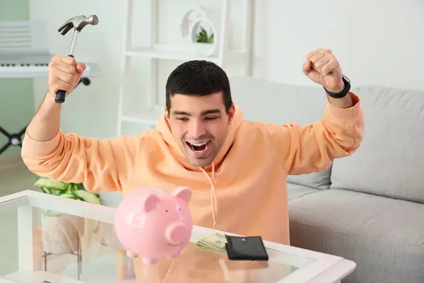 Happy young man with hammer breaking piggy bank at home