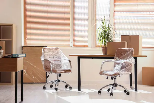 Interior of light office with chairs wrapped in stretch film and cardboard boxes on moving day