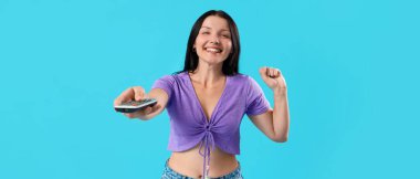Joyful young woman with remote control watching TV on blue background clipart