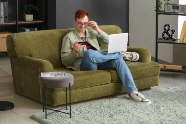 İnternetten alışveriş yaparken koltukta oturan, kredi kartı ve modern dizüstü bilgisayarı olan yakışıklı bir genç.