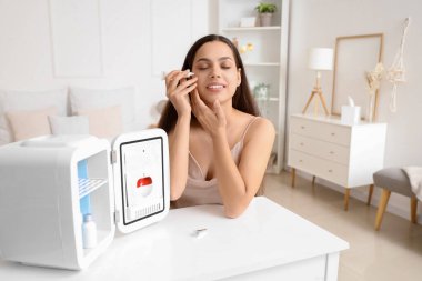 Kozmetik ürünleri için küçük bir buzdolabı olan güzel genç bir kadın yatak odasında krem sürüyor.