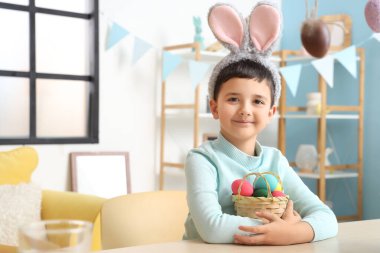 Tavşan kulaklı ve sepetinde Paskalya yumurtaları olan sevimli küçük çocuk dekore edilmiş odada masada oturuyor.