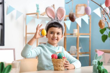 Tavşan kulaklı ve sepetinde Paskalya yumurtaları olan sevimli küçük çocuk dekore edilmiş odada masada oturuyor.