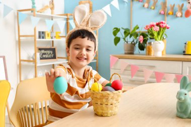 Tavşan kulaklı ve sepetinde Paskalya yumurtaları olan sevimli küçük çocuk dekore edilmiş odada masada oturuyor.