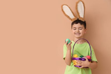 Tavşan kulaklı şirin çocuk elinde hasır sepetle bej arka planda Paskalya yumurtaları tutuyor.