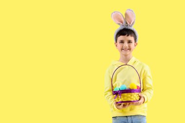 Tavşan kulaklı şirin bir çocuk elinde sepet ve sarı arka planda Paskalya yumurtaları var.