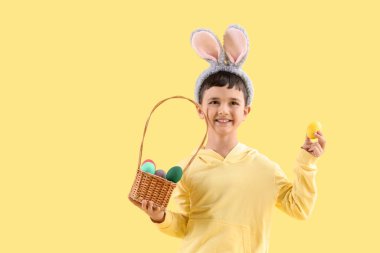 Tavşan kulaklı şirin bir çocuk elinde sepet ve sarı arka planda Paskalya yumurtaları var.
