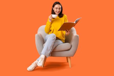 Elinde çay fincanı olan genç bir kadın turuncu arka planda koltukta kitap okuyor.