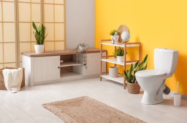 Ev bitkileri ve turuncu duvarın yanında seramik klozeti olan şık bir banyo.