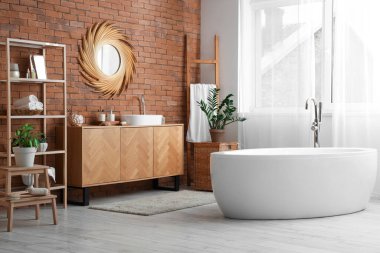 Tuğla duvarda asılı yuvarlak ayna, beyaz küvet, banyonun içinde cilt bakım ürünleri olan ahşap raf ünitesi.