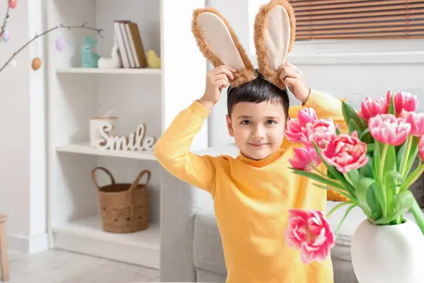 Cute little boy in bunny ears sitting at table with tulip flowers at home. Easter celebration