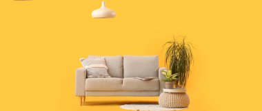 Sarı arka planda yastıklı, puflu, çiçekli ve tavan lambalı rahat gri kanepe.