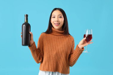 Mavi arka planda cam ve kırmızı şarap şişesiyle güzel bir Asyalı kadın.