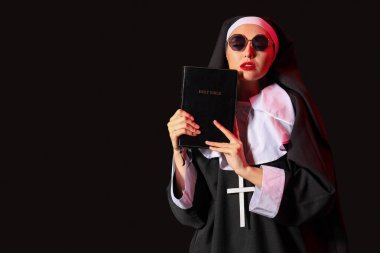 Karanlık arka planda kutsal kitapla güneş gözlüklü yaramaz rahibe.