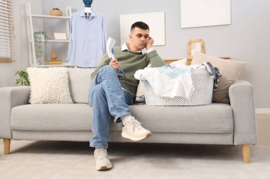 Buharlı tenceresi ve çamaşır dolu sepeti olan yorgun genç adam evdeki kanepede oturuyor.
