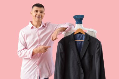 Mutlu genç adam pembe arka planda modern bir vapura asılı şık bir ceketi işaret ediyor.