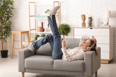 Kulaklıklı, cep telefonlu, rahat bir kanepeye uzanmış ve evdeki oturma odasında müzik dinleyen güzel bir kadın.