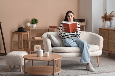 Evdeki oturma odasında rahat bir koltukta oturup kitap okuyan güzel genç bir kadın.