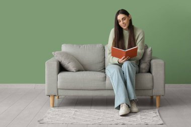 Dizüstü bilgisayarı olan genç ve güzel bir kadın rahat koltukta oturuyor ve yeşil duvarın yanında kitap okuyor.