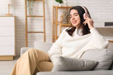 Kulaklıklı, güzel, genç ve mutlu bir kadın rahat bir koltukta oturup evde müzik dinliyor.