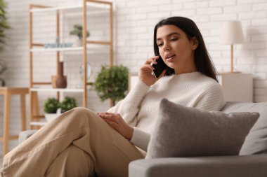 Evdeki oturma odasında rahat bir koltukta oturan ve cep telefonuyla konuşan güzel genç bir kadın.