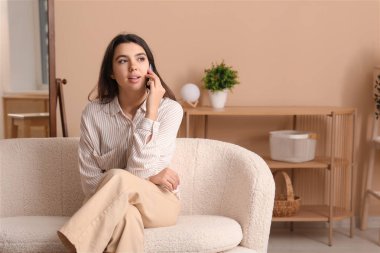 Evdeki oturma odasında rahat bir koltukta oturan ve cep telefonuyla konuşan güzel genç bir kadın.