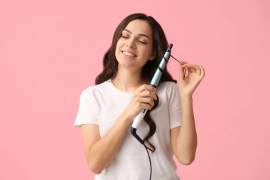 Beautiful young woman with curling iron on pink background