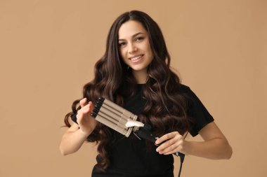 Beautiful young woman with curling iron on beige background