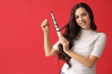 Beautiful young woman with curling iron on red background