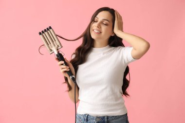 Beautiful young woman with curling iron on pink background