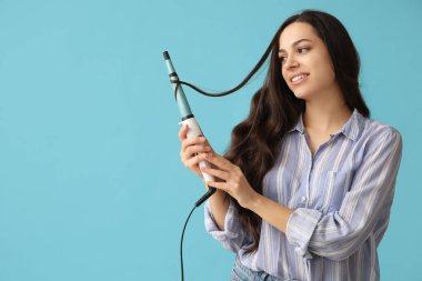 Beautiful young woman with curling iron on blue background