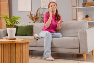 Gülümseyen kızıl saçlı kadın evdeki koltukta cep telefonuyla konuşuyor.