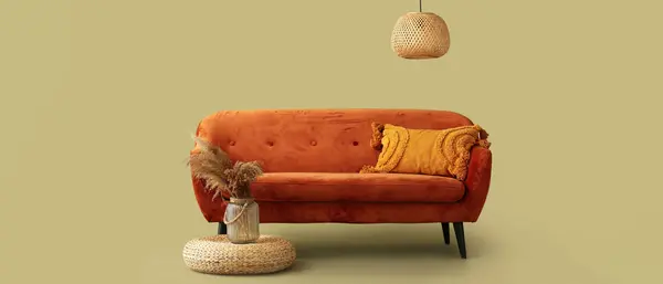 Cozy brown sofa with cushion, wicker pouf and ceiling lamp on green background
