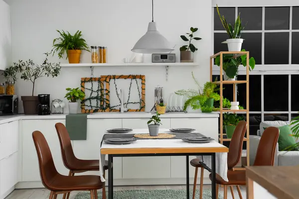 Modern mutfağın içinde yeşil bitkiler, yemek masası ve tezgahlar var.