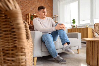 Evdeki kanepede diz ağrısı çeken genç bir adam.