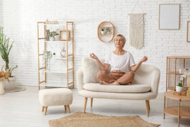 Olgun bir kadın evde kanepede otururken meditasyon yapıyor.