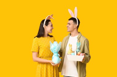 Paskalya tavşanı kulaklı mutlu genç çift, sarı arka planda sepet ve hediye.