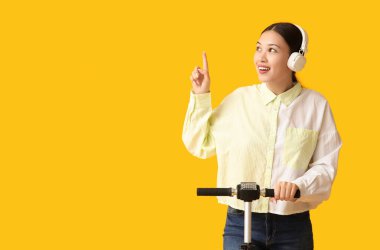 Kulaklıklı, modern elektrikli scooterlı güzel Asyalı kadın sarı arka plandaki bir şeyi işaret ediyor.