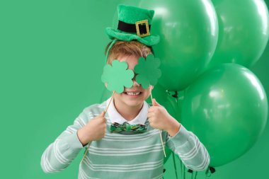 Yeşil arka planda leprikon şapkalı, yoncalı ve balonlu şirin bir çocuk. Aziz Patrick Günü kutlaması