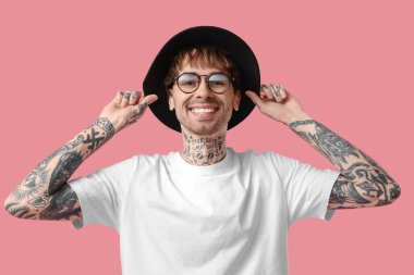 Pembe arka planda şapka ve gözlük takan dövmeli genç bir adam.