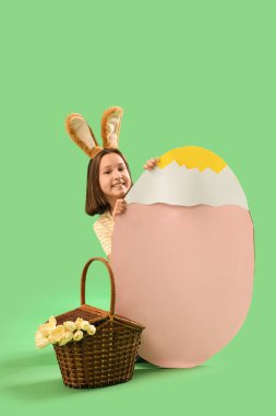 Paskalya tavşanı kulaklı şirin küçük kız, yeşil arka planda büyük kağıt yumurta ve sepet.