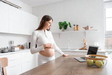 Mutfaktaki masada dizüstü bilgisayarı olan hamile bir kadın.
